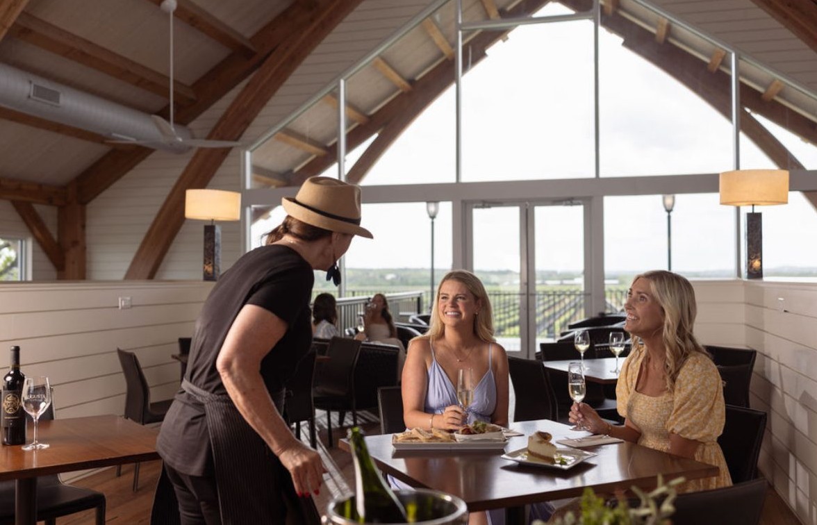Two guests smiling at a tasting table while a winery host pours